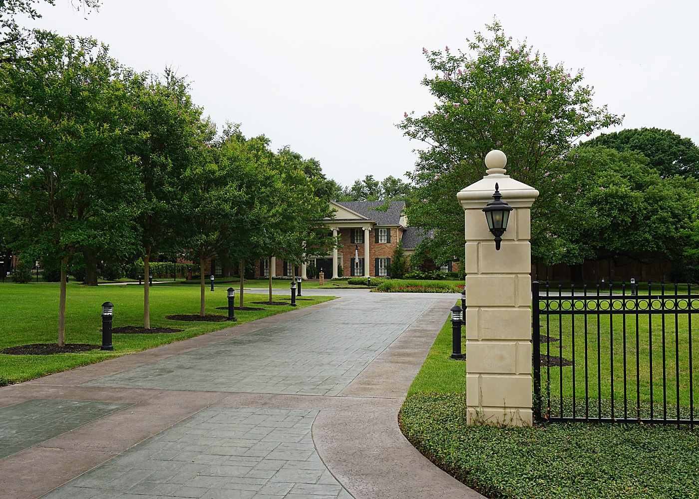 A landscaped entrance drive with stone gate posts and iron railings leading into a wooded residential property — representative imagery, not a specific Texas Pride job